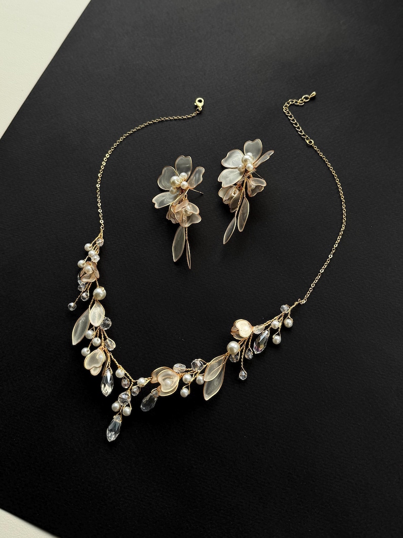 K&ouml;nnte beinhalten: Ein zartes, goldfarbenes Halsketten- und Ohrring-Set. Die Halskette und die Ohrringe verf&uuml;gen &uuml;ber florale und blattf&ouml;rmige Akzente mit Kunstperlen und klaren Kristallverzierungen. Der Schmuck wird auf einer schwarzen Oberfl&auml;che pr&auml;sentiert.