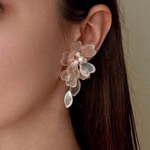 K&ouml;nnte beinhalten: Nahaufnahme eines floralen Ohrrings. Der Ohrring hat mehrere durchscheinende Bl&uuml;tenbl&auml;tter in Rosa- und Wei&szlig;t&ouml;nen mit einem kleinen Perlenakzent in der Mitte. Der Ohrring ist an einem goldfarbenen Stecker befestigt.