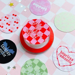 May include: A collection of round stickers with various designs and text. One sticker has a red and pink checkerboard pattern with the words "Hot Girls Read." Other stickers feature cherries, a heart, and the words "fantasy girlie," "emotional support kindle," and "Do Not Disturb."