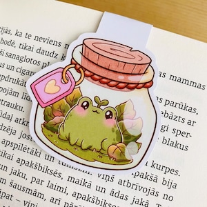 Op de afbeelding: Een grillige boekenlegger met een cartoonkikker in een pot. De kikker is groen met grote ogen en een kleine spruit. De pot heeft een kurken deksel en een roze label met een hart. De boekenlegger ligt op een boek.