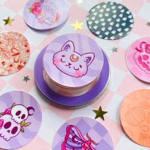 Op de afbeelding: Een verzameling ronde stickers en een kleine container met een paarse deksel. De stickers hebben verschillende ontwerpen, waaronder een kat met een maan, schedels met rozen, een mot, een pompoen en een boek. Het kleurenpalet omvat paars, roze en oranje.