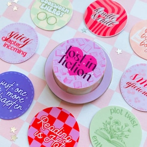 Op de afbeelding: Een verzameling ronde stickers met boeken-thema's en teksten. De centrale sticker toont "lost in fiction" in een hart. Andere stickers tonen "Bookworm Energy" en "Reading is Hat". Ze liggen op een roze en witte geruite achtergrond.