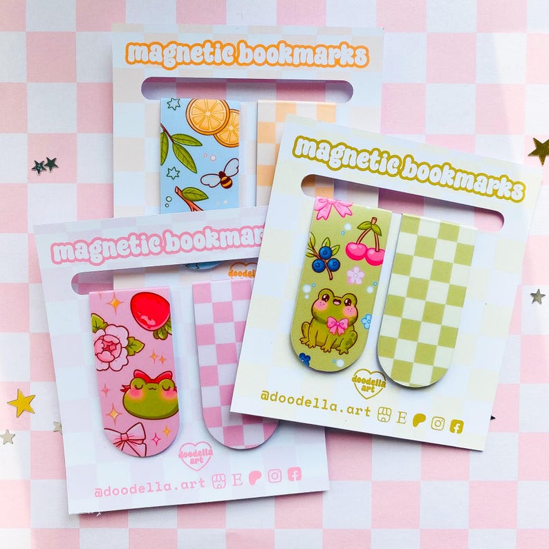 May include: Several magnetic bookmarks are displayed, each with unique designs. One features a frog with a bow, another has lemons and a bee, and a third showcases cherries and blueberries. The packaging reads "magnetic bookmarks" in a pink font.
