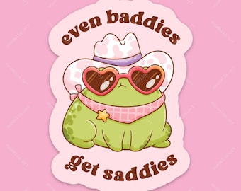 Op de afbeelding: Een gestanste sticker met een groene kikker die een wit met roze cowboyhoed met koeienprint, hartvormige zonnebril en een roze geblokte bandana met een gele ster draagt. De tekst luidt even baddies get saddies in bruine letters.