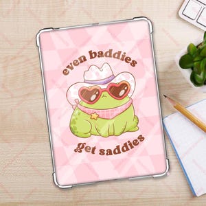 May include: A pink and white checkered background with a cartoon frog wearing a cowboy hat and heart-shaped sunglasses. The text "even baddies get saddies" is written in brown letters.