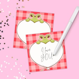May include: Two square cards with a red and white gingham pattern. Each card features a green frog holding a white heart with the words "I love you" written on one. A white stylus pen rests on the cards against a pink background.