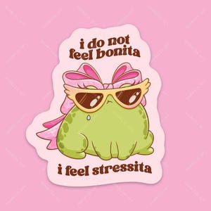 May include: A die-cut sticker featuring a green frog wearing large cat-eye sunglasses and a pink bow. The text above reads 'i do not feel bonita' and the text below reads 'i feel stressita' in brown lettering against a light pink background.