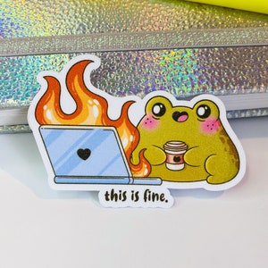 Op de afbeelding: Een gestanste sticker met een groene kikker met roze wangen die een koffiekopje vasthoudt naast een blauwe laptop die in oranje vlammen staat. De sticker bevat de tekst this is fine. in zwarte kleine letters aan de onderkant.