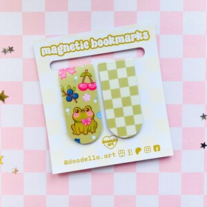 May include: Two magnetic bookmarks on a white card with the text "magnetic bookmarks". One bookmark features a cartoon frog, cherries, and blueberries. The other has a green and white checkered pattern. The card is on a pink and white checkered surface.