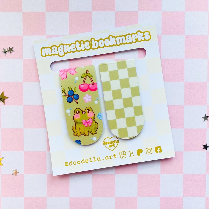 May include: Two magnetic bookmarks on a white card with the text "magnetic bookmarks". One bookmark features a cartoon frog, cherries, and blueberries. The other has a green and white checkered pattern. The card is on a pink and white checkered surface.