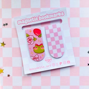 May include: Two magnetic bookmarks on a pink and white checkered background. One bookmark features a frog, flowers, and a bow. The other has a pink and white checkerboard pattern. The words "magnetic bookmarks" are at the top.