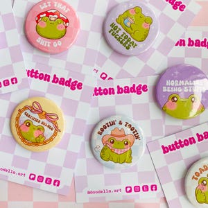 May include: A selection of button badges with frog designs and humorous text. The badges are circular and feature illustrations of frogs in different scenarios, such as under a mushroom or wearing a cowboy hat. Text includes phrases like "Not Today Fuckers" and "Rootin' & Tootin'."