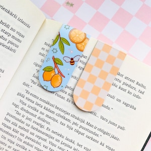 May include: Two decorative bookmarks. One features a light blue background with illustrations of lemons, leaves, and a bee. The other has an orange and white checkered pattern. Both are placed on an open book.