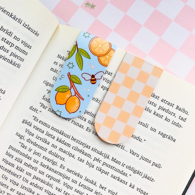 May include: Two decorative bookmarks. One features a light blue background with illustrations of lemons, leaves, and a bee. The other has an orange and white checkered pattern. Both are placed on an open book.