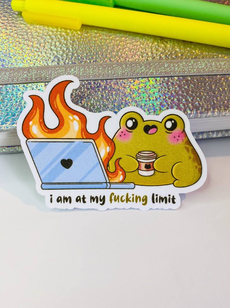 Op de afbeelding: Een gestanste sticker met een groene kikker met roze wangen die een koffiebeker vasthoudt naast een laptop die in oranje vlammen opgaat. De tekst onder de kikker luidt "i am at my fucking limit" in zwarte, kleine, schreefloze letters.