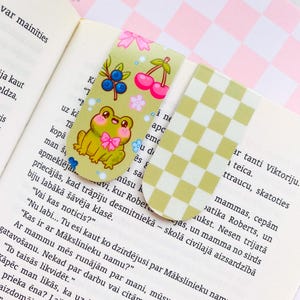 May include: Two decorative bookmarks. One features a cartoon frog with a pink bow, blueberries, cherries, and flowers on a light green background. The other has a yellow and white checkered pattern.