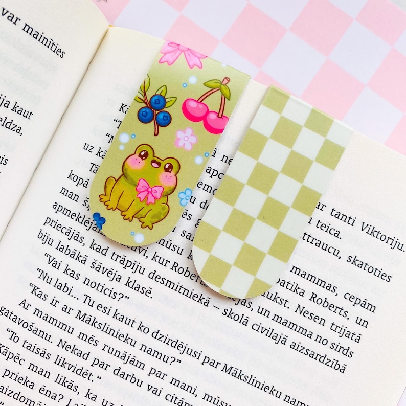 May include: Two decorative bookmarks. One features a cartoon frog with a pink bow, blueberries, cherries, and flowers on a light green background. The other has a yellow and white checkered pattern.