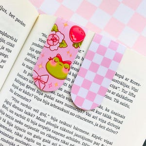 May include: Two decorative bookmarks. One pink bookmark features a green frog with a red bow, flowers, and a red strawberry. The other bookmark has a pink and white checkered pattern.