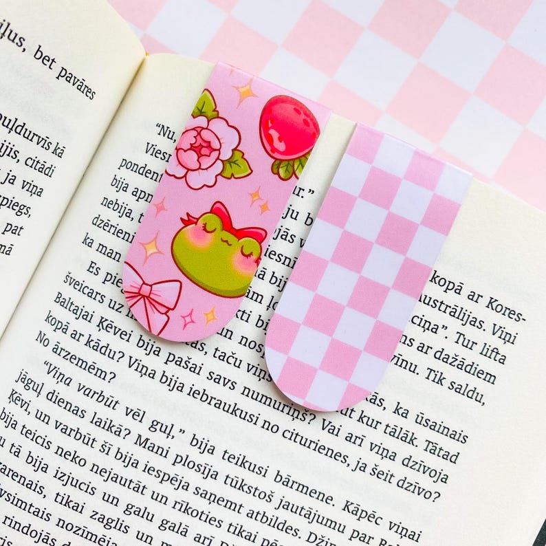 May include: Two decorative bookmarks. One pink bookmark features a green frog with a red bow, flowers, and a red strawberry. The other bookmark has a pink and white checkered pattern.