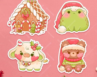 Op de afbeelding: Vier stickers met een peperkoekhuisje met zuurstokken, een groene kikker met een rode kerstmuts, een crèmekleurige koe met hulst en een belletje, en een bruine beer met een rode kerstmuts en een groene trui op een roze achtergrond.