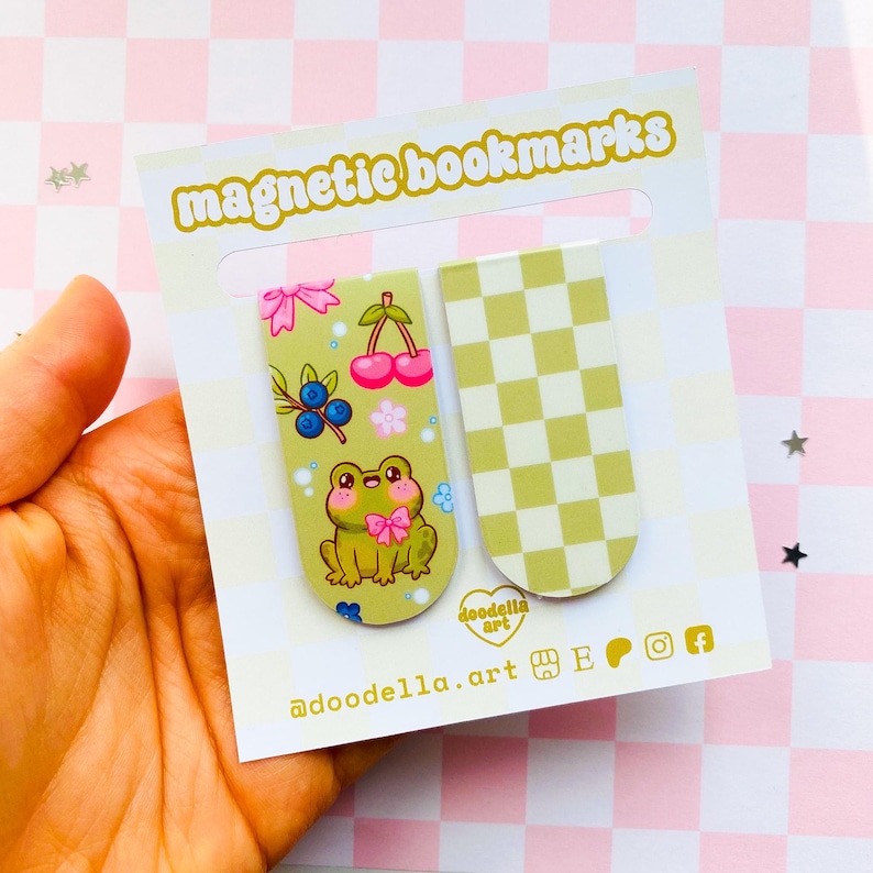 May include: Magnetic bookmarks featuring a frog, cherries, and blueberries on a light green background. The second bookmark has a green and white checkered pattern. The card reads "magnetic bookmarks" and "@doodella.art".