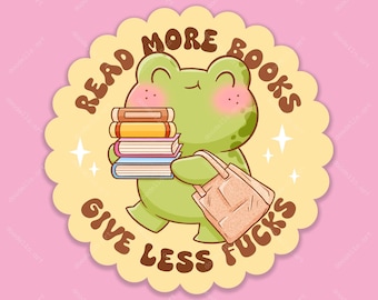 Op de afbeelding: Een ronde sticker met een geschulpte rand met een groene kikker met roze wangen die een stapel van vier boeken en een beige tas vasthoudt. De tekst READ MORE BOOKS GIVE LESS FUCKS staat in bruine letters op een lichtgele achtergrond.