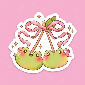 May include: A die-cut sticker featuring two light green, round frog characters with rosy cheeks, connected by brown stems to a single pink ribbon tied in a bow. The design includes a small green leaf and several four-pointed yellow stars.