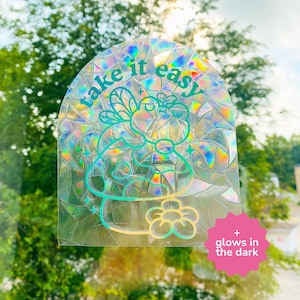 May include: Holographic sticker with the words "take it easy" in teal script. The sticker features a whimsical design of a creature and a flower. A pink starburst graphic indicates the sticker glows in the dark.