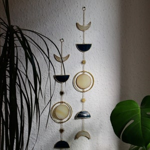 May include: Two celestial-themed wall hangings with gold-colored chains and accents. Each features crescent moons, circular shapes, and starbursts in cream, navy, and gold. The decor is hanging on a textured white wall, with green plants in the foreground.
