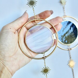 May include: A celestial-themed hanging decoration featuring gold-colored metal rings and sunburst charms. The circular glass inserts display a mix of colors, including white, pink, and blue. The design is reminiscent of the moon phases.