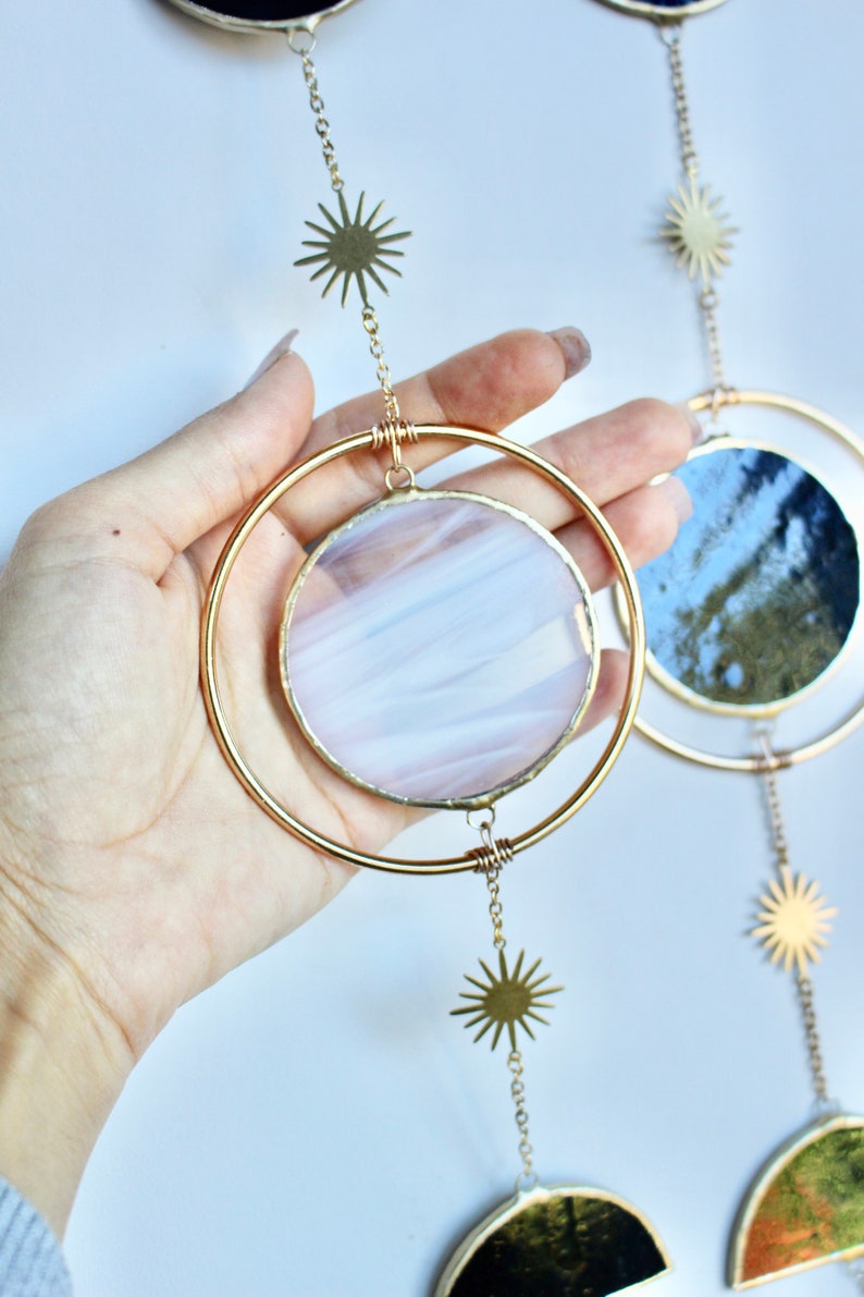 May include: A celestial-themed hanging decoration featuring gold-colored metal rings and sunburst charms. The circular glass inserts display a mix of colors, including white, pink, and blue. The design is reminiscent of the moon phases.