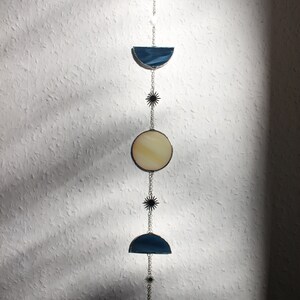 May include: A celestial-themed wall hanging featuring phases of the moon. The design includes crescent, semi-circle, and circular shapes in cream and blue stained glass, connected by silver chains and star accents. The piece is suspended against a textured white wall.