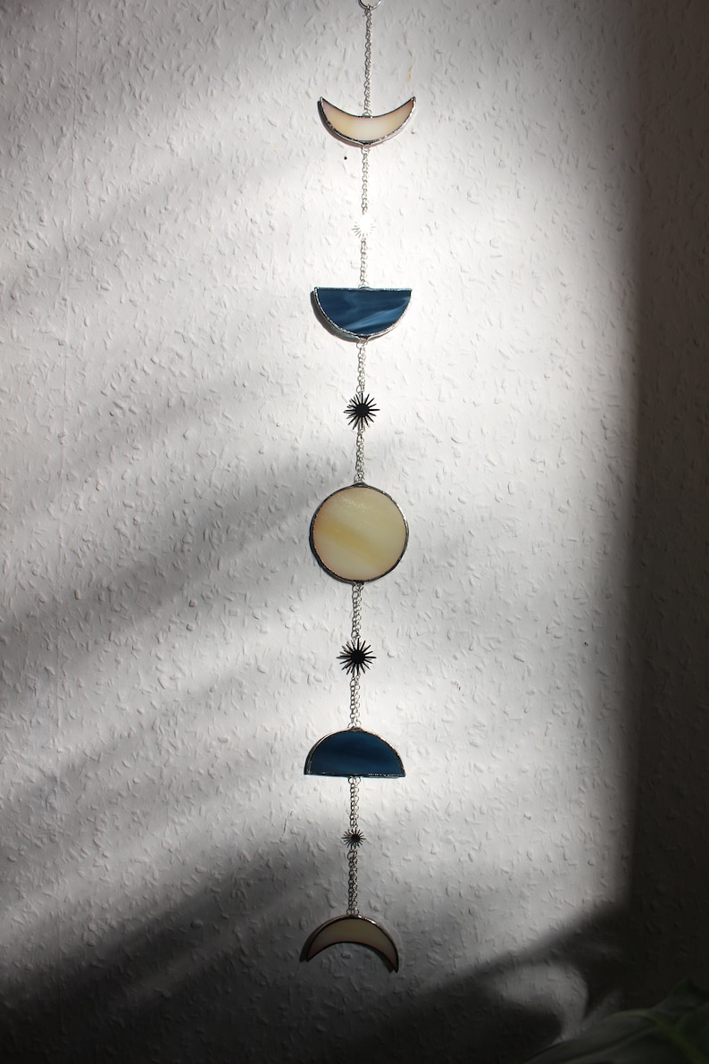 May include: A celestial-themed wall hanging featuring phases of the moon. The design includes crescent, semi-circle, and circular shapes in cream and blue stained glass, connected by silver chains and star accents. The piece is suspended against a textured white wall.