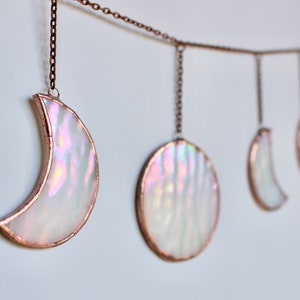 Op de afbeelding: Een decoratieve wandhanger met iriserende maanfasen. Elke fase is omlijnd met koperkleurig metaal en hangt aan een delicate ketting. De fasen variëren van een halve maan tot een volle maan.