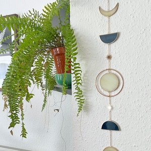 May include: A celestial-themed wall hanging featuring a series of moon phases and stars. The design incorporates a crescent moon, a half-moon shape, a full moon circle, and star accents, all connected by delicate gold chains. The moon shapes have a cream and navy blue color scheme.