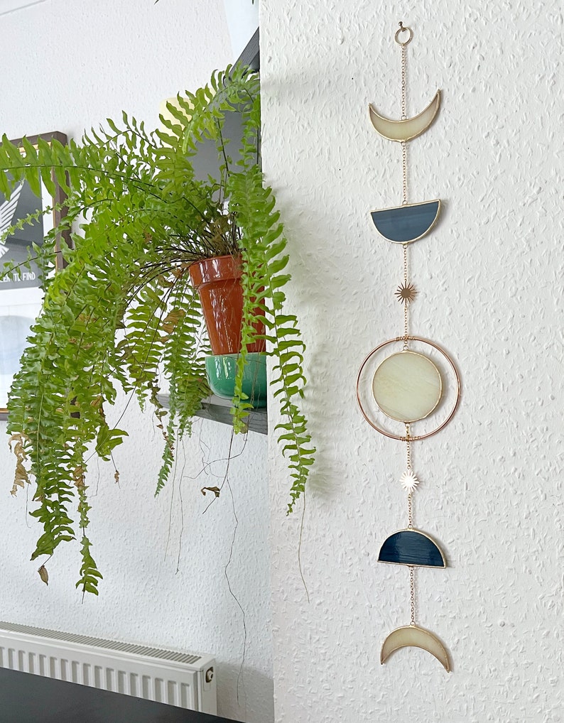 May include: A celestial-themed wall hanging featuring a series of moon phases and stars. The design incorporates a crescent moon, a half-moon shape, a full moon circle, and star accents, all connected by delicate gold chains. The moon shapes have a cream and navy blue color scheme.