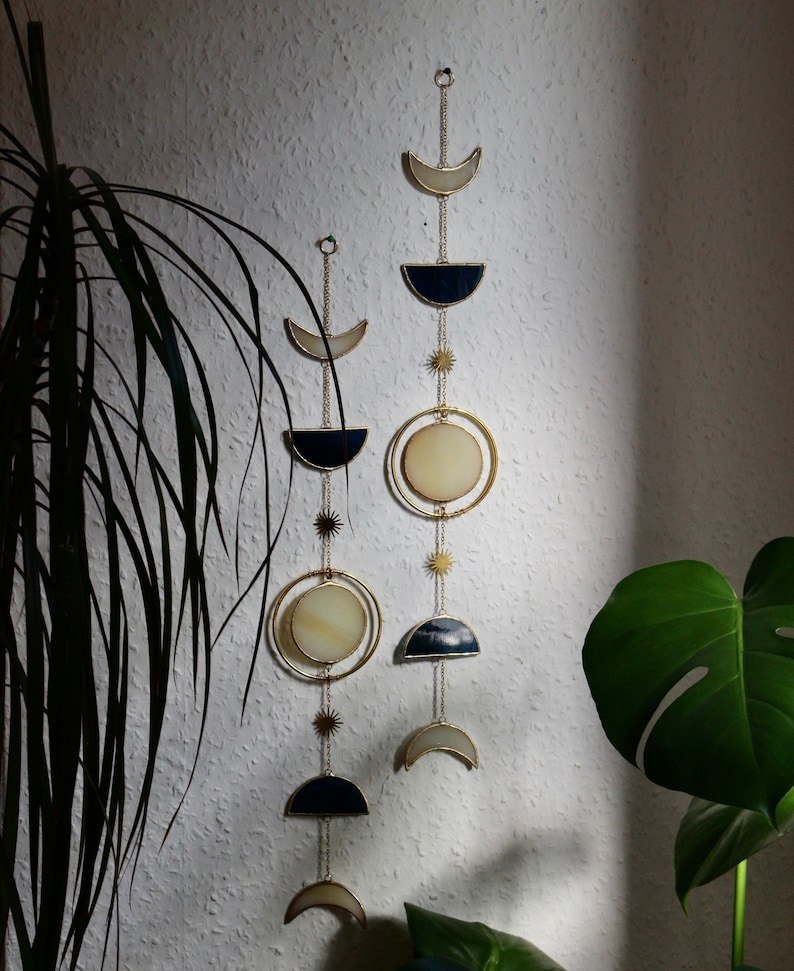 May include: Two celestial-themed wall hangings with gold-colored chains and accents. Each features crescent moons, circular shapes, and starbursts in cream, navy, and gold. The decor is hanging on a textured white wall, with green plants in the foreground.