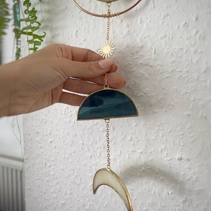 May include: A celestial-themed stained glass suncatcher. It features a gold-colored metal frame with a crescent moon, sun, and a half-circle design in shades of blue and white. The suncatcher is suspended by a delicate chain.