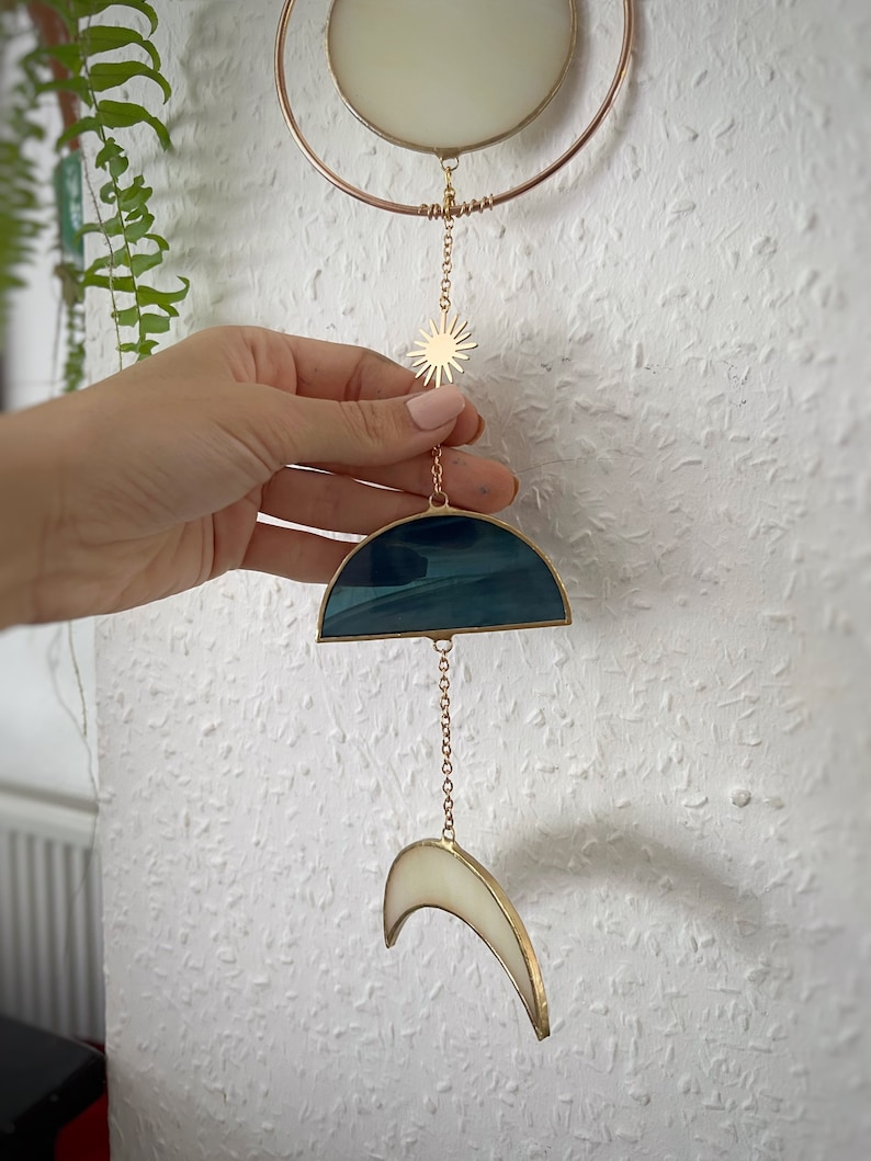 May include: A celestial-themed stained glass suncatcher. It features a gold-colored metal frame with a crescent moon, sun, and a half-circle design in shades of blue and white. The suncatcher is suspended by a delicate chain.