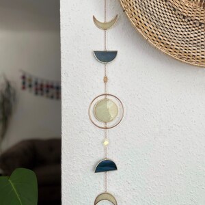 May include: A celestial-themed wall hanging featuring phases of the moon. The design includes a crescent, a half-moon, and a full moon, all in shades of cream and navy blue, connected by delicate chains. A sunburst charm adds a touch of sparkle.