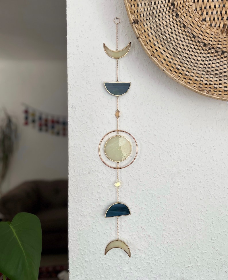 May include: A celestial-themed wall hanging featuring phases of the moon. The design includes a crescent, a half-moon, and a full moon, all in shades of cream and navy blue, connected by delicate chains. A sunburst charm adds a touch of sparkle.