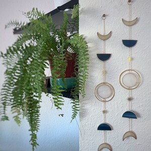 May include: Two celestial-themed wall hangings with gold-colored chains and accents. Each features a crescent moon, a half-circle, a circle, and star-shaped charms. The wall hangings are in front of a white textured wall. A potted fern is on a shelf to the left.