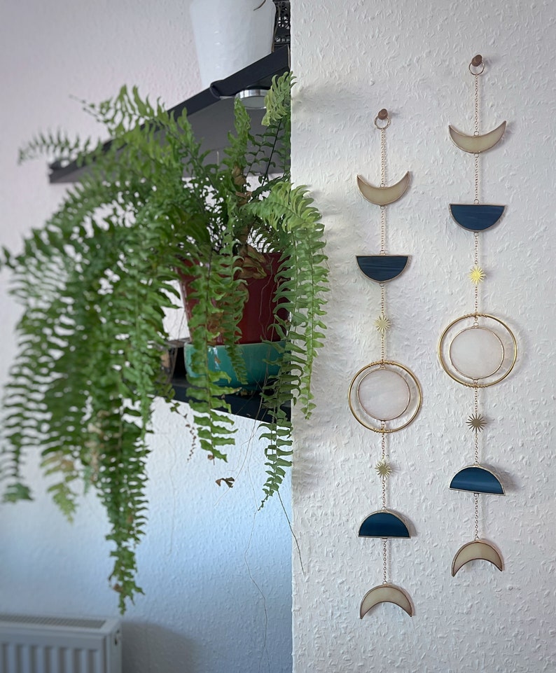 May include: Two celestial-themed wall hangings with gold-colored chains and accents. Each features a crescent moon, a half-circle, a circle, and star-shaped charms. The wall hangings are in front of a white textured wall. A potted fern is on a shelf to the left.