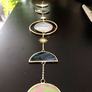 May include: A celestial-themed wall hanging featuring a series of gold-toned metal shapes and stained glass elements. The design includes crescent moons, a large oval, and starburst accents, creating a decorative and artistic piece.