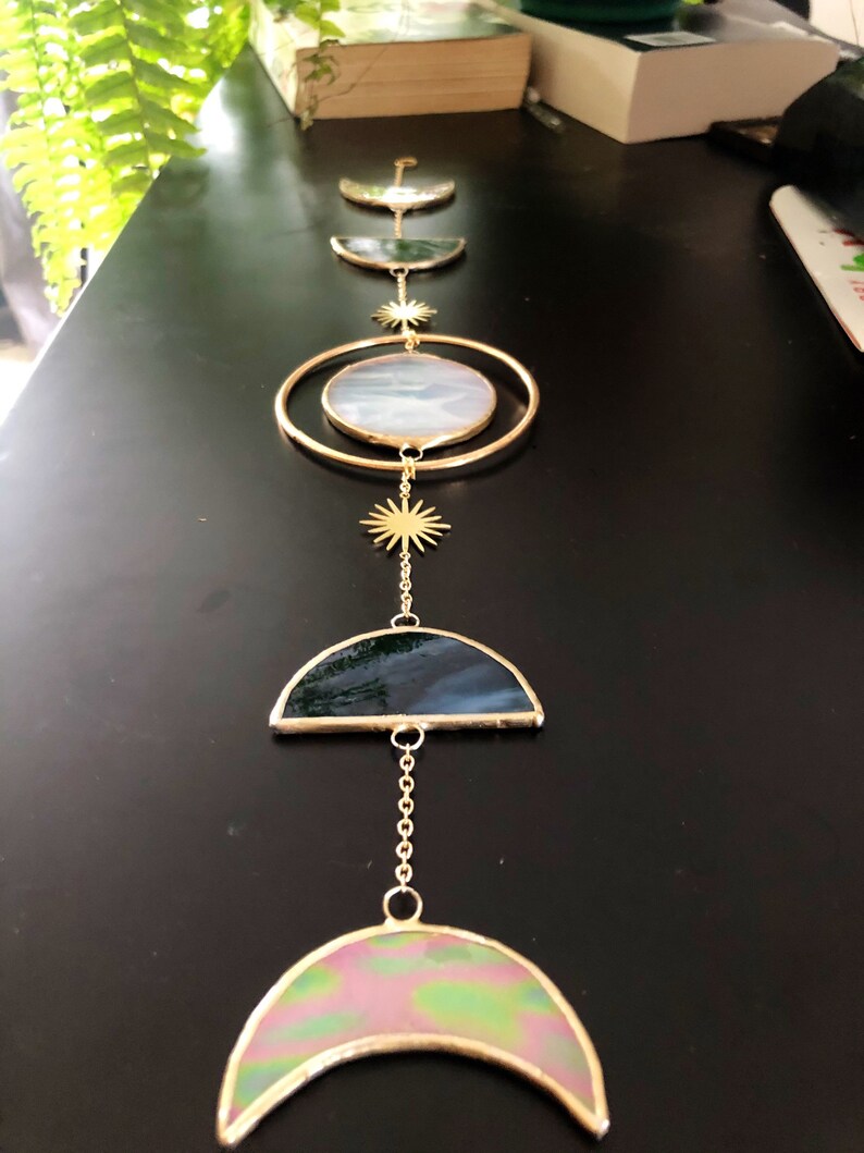 May include: A celestial-themed wall hanging featuring a series of gold-toned metal shapes and stained glass elements. The design includes crescent moons, a large oval, and starburst accents, creating a decorative and artistic piece.