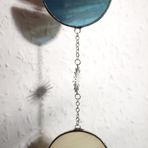 May include: A stained glass suncatcher with a half-moon shape in teal and a circular shape in cream, connected by a silver chain. A star-shaped charm is also on the chain. The suncatcher is held by a hand.