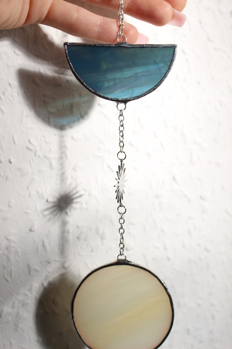 May include: A stained glass suncatcher with a half-moon shape in teal and a circular shape in cream, connected by a silver chain. A star-shaped charm is also on the chain. The suncatcher is held by a hand.