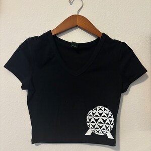 Op de afbeelding: Zwart getailleerd T-shirt met een witte afbeelding van de Epcot Spaceship Earth.