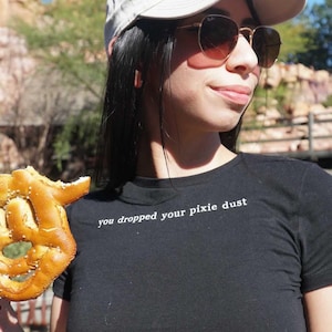 May include: A woman wearing a black t-shirt with the text "you dropped your pixie dust" in white lettering. She is holding a pretzel in her left hand and wearing a white baseball cap and brown sunglasses.