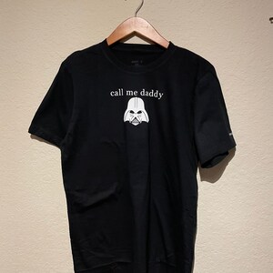 Op de afbeelding: Zwart T-shirt met een witte afbeelding van Darth Vaders helm en de tekst "call me daddy".